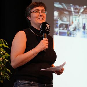 Frau mit Mikrofon und Zettel in der Hand vor großer Leinwand sprechend 