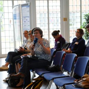 Frau mit Mikrofon auf Stuhl sitzend neben anderen Personen in Stuhlreihe sitzend 